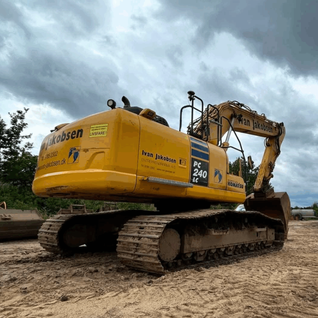 Yellow hydraulic excavator with tracks sits on a dirt construction site under a cloudy sky, labeled Ivan Jakobsen/KOMATSU PC240LC.