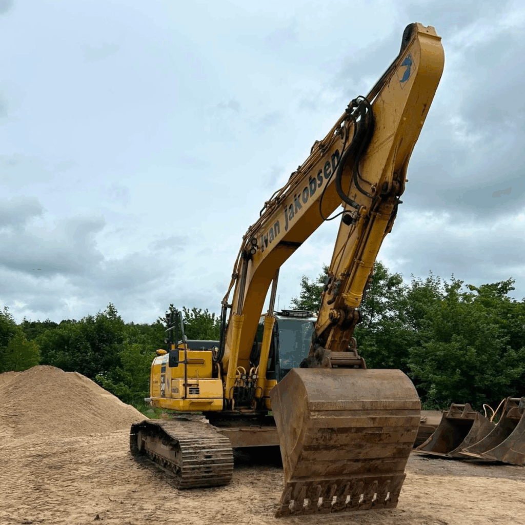 Yellow excavator with extended arm and bucket on a dirt construction site, a pile of soil nearby and trees in the background